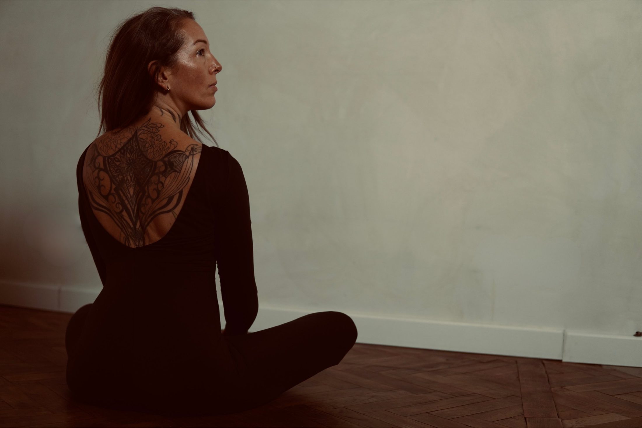 Chiara sitzt im Schneidersitz am Boden, Blick zur Seite, Tattoo am Rücken