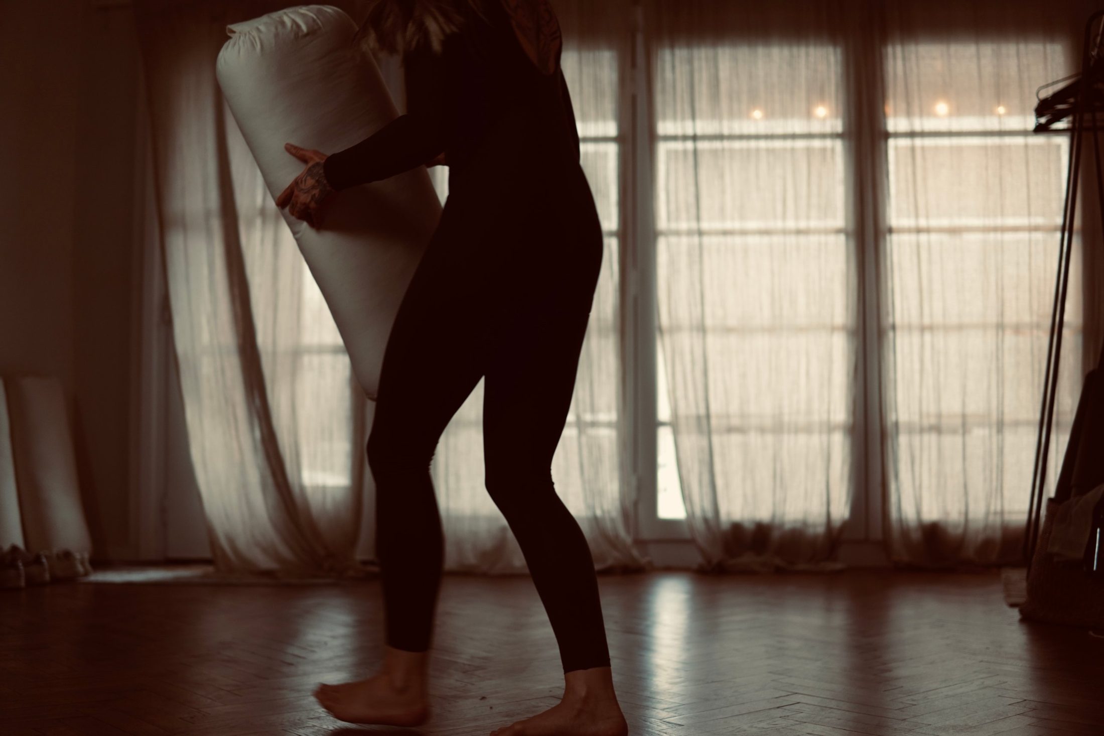 Chiara trägt eine Yogarolle durch den raum., hinter ihr Fenster mit Leinenvorhängen